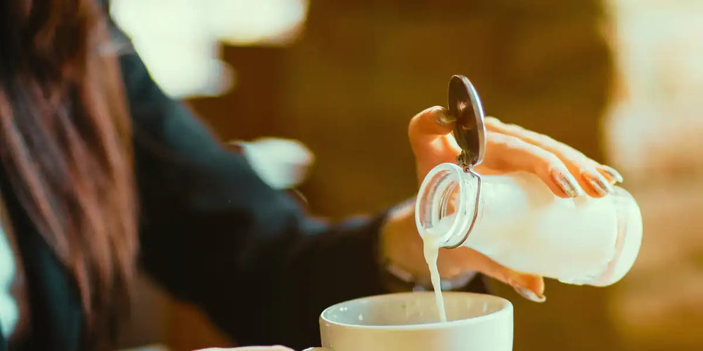 Leite sendo servido de uma jarra de vidro em uma xícara branca de café, ilustrando o ajuste da proporção entre café e leite.