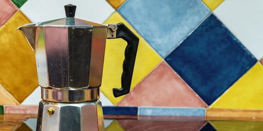 Cafeteira italiana Moka clássica em alumínio sobre bancada, com fundo de azulejos coloridos