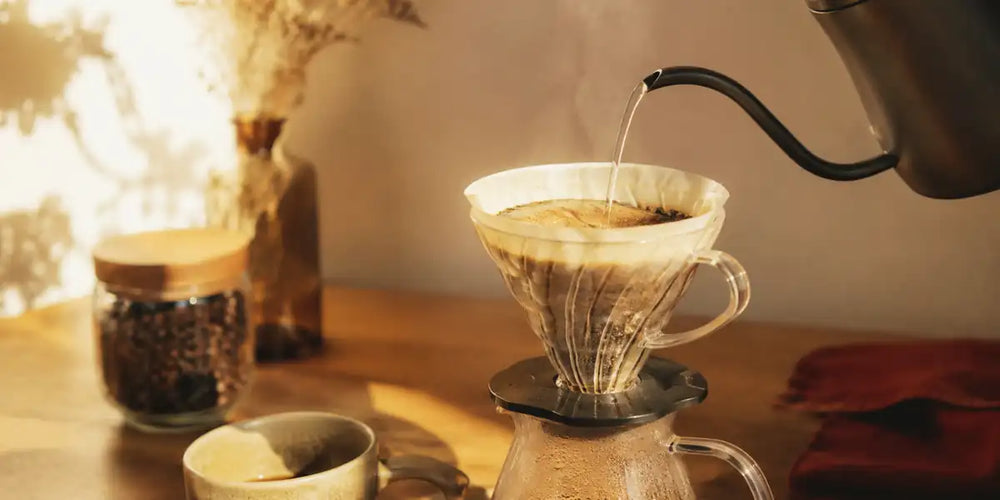 Chaleira pescoço de ganso despejando fio de água em coador V60 sobre bancada de madeira