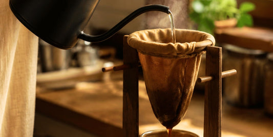 Água quente sendo despejada por um coador de pano tradicional, com café fresco pingando em um bule de cerâmica.