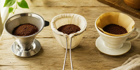 Três coadores de café lado a lado: filtro de inox, coador de pano e porta-filtro com papel, cada um com café moído, sobre mesa de madeira rústica