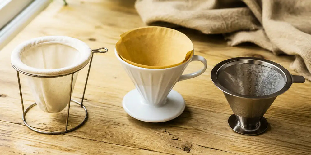 Três tipos de coador de café sobre bancada de madeira: pano, papel e inox lado a lado