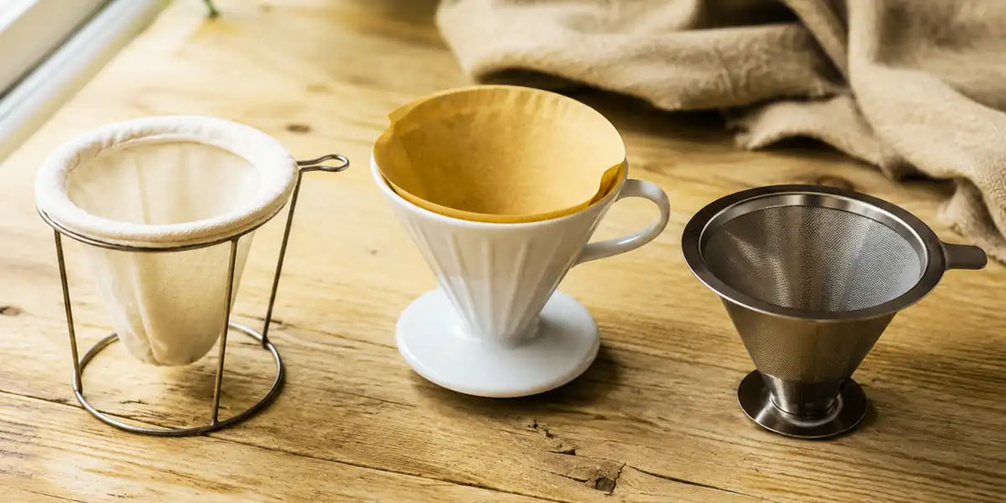 Três tipos de coador de café sobre bancada de madeira: pano, papel e inox lado a lado