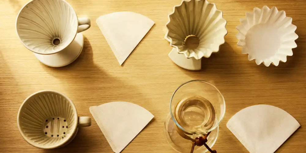 Quatro coadores de café diferentes, cada um ao lado do seu filtro de papel correspondente, sobre superfície de madeira clara.