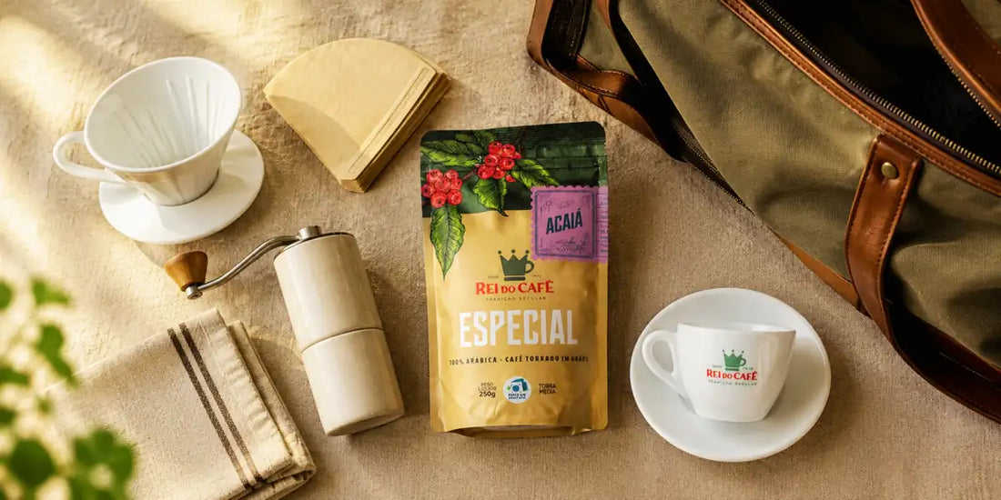 Kit de café para viagem visto de cima sobre linho, com coador, filtros, moedor manual, pacote Rei do Café Especial Acaiá e xícara ao lado de mala aberta
