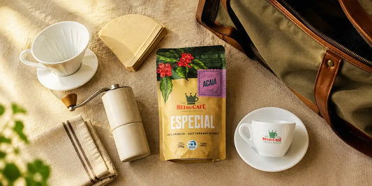 Kit de café para viagem visto de cima sobre linho, com coador, filtros, moedor manual, pacote Rei do Café Especial Acaiá e xícara ao lado de mala aberta