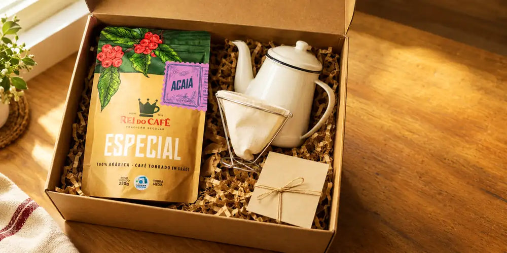 Caixa de presente aberta com pacote de café especial Rei do Café, bule esmaltado, coador de pano e cartão sobre mesa de madeira