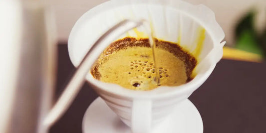 Água quente despejada em coador cônico branco, formando bloom sobre o café moído durante a extração