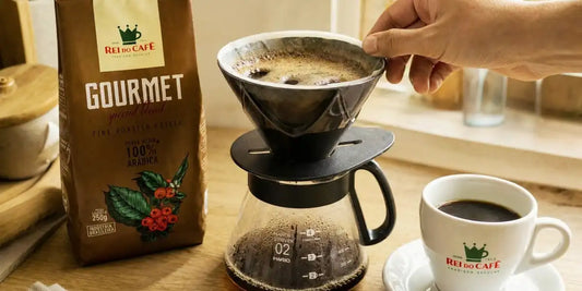 Mão girando suavemente o coador durante a pré-infusão do café coado, ao lado do pacote Rei do Café Gourmet e de uma xícara