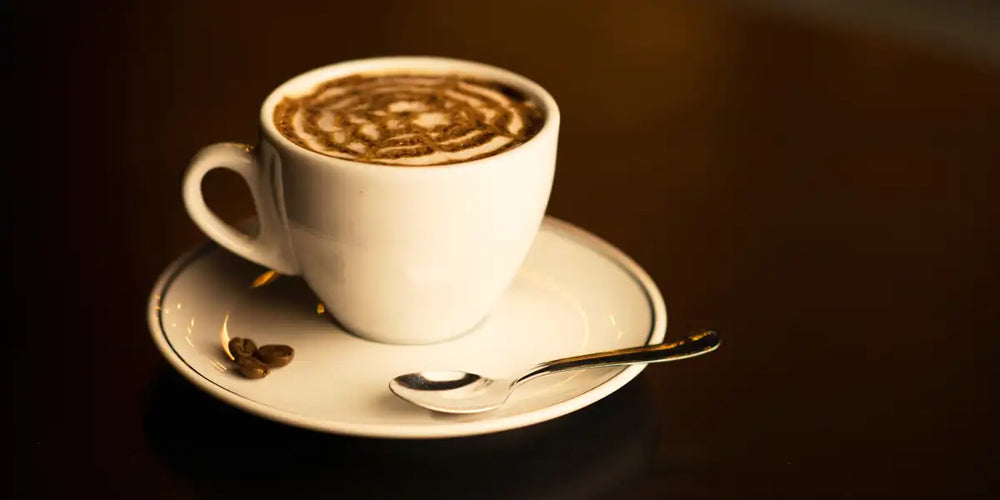 Xícara branca de mocaccino caseiro com desenho de cacau em pó sobre a espuma, pires com grãos de café e colher