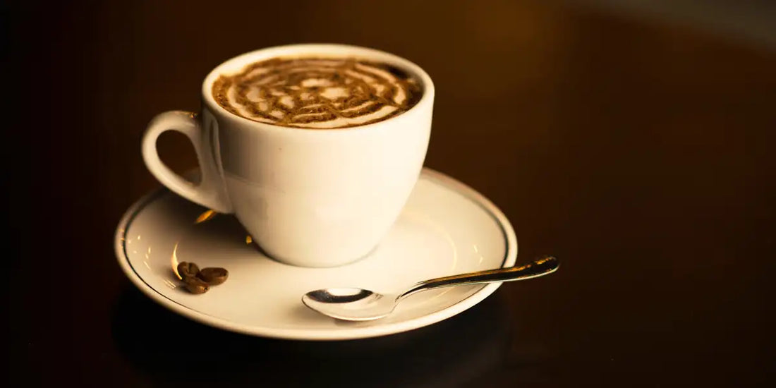 Xícara branca de mocaccino caseiro com desenho de cacau em pó sobre a espuma, pires com grãos de café e colher