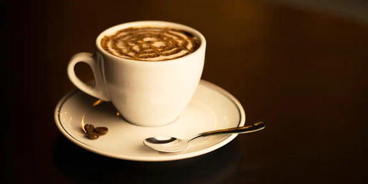 Xícara branca de mocaccino caseiro com desenho de cacau em pó sobre a espuma, pires com grãos de café e colher