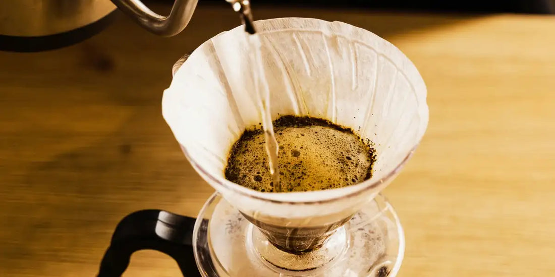 Água quente sendo despejada de chaleira sobre filtro de papel branco em coador V60 de vidro, com café em bloom