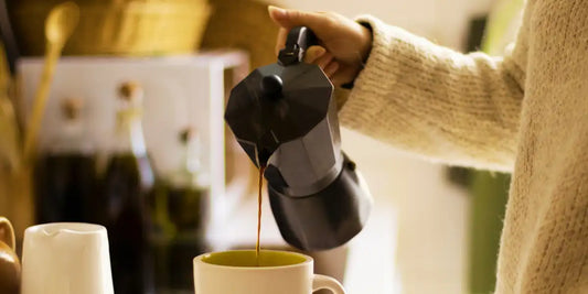 Pessoa servindo café de uma cafeteira italiana em uma caneca de cerâmica sobre a bancada.