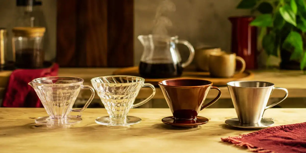 Coadores de café em plástico, vidro, cerâmica e aço inoxidável lado a lado sobre bancada de madeira.
