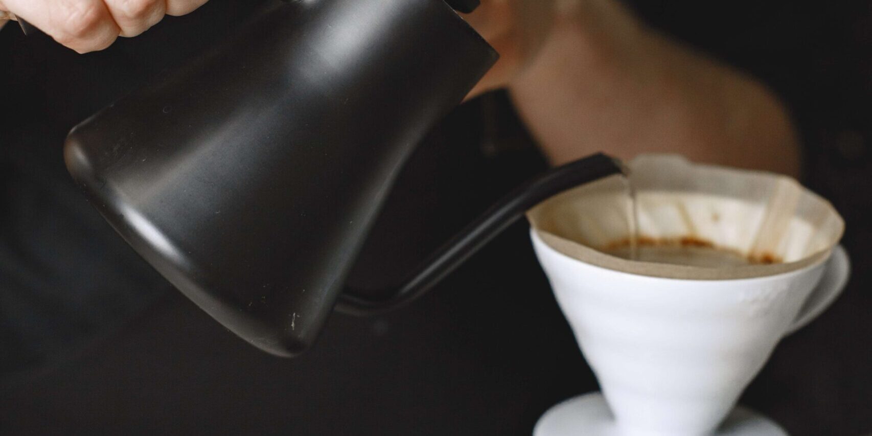 imagem de duas mãos derramando água de um bule preto em um filtro de café branco que está em cima de uma xícara transparente