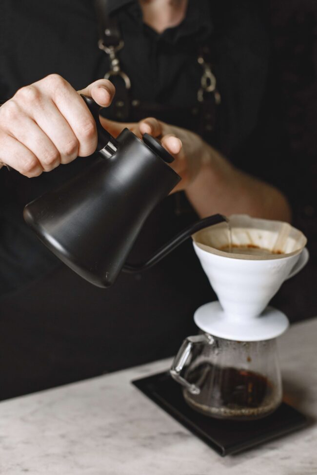 imagem de duas mãos derramando água de um bule preto em um filtro de café branco que está em cima de uma xícara transparente