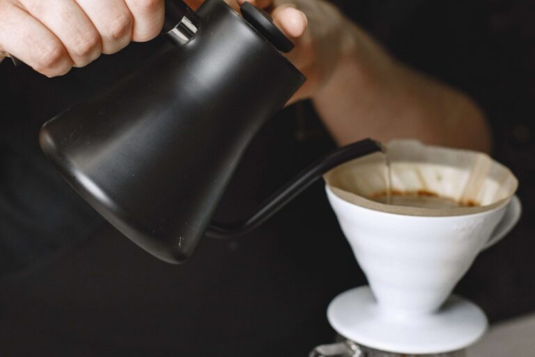 imagem de duas mãos derramando água de um bule preto em um filtro de café branco que está em cima de uma xícara transparente