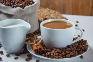 imagem de uma xícara branca com café e ao redor grãos de café e canela em pau sobre uma mesa branca com alguns grãos de café ao redor, uma xícara de leite ao lado e uma saca de café em grãos atrás