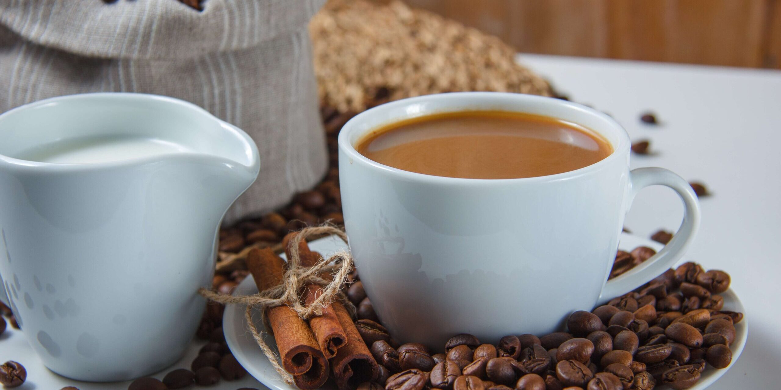 imagem de uma xícara branca com café e ao redor grãos de café e canela em pau sobre uma mesa branca com alguns grãos de café ao redor, uma xícara de leite ao lado e uma saca de café em grãos atrás