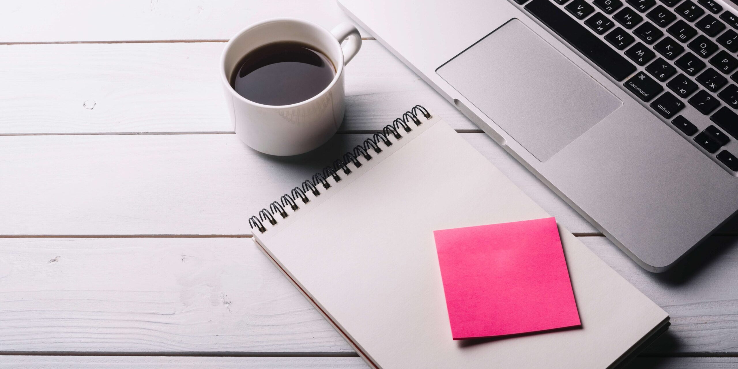 imagem de uma xícara branca com café preto, em cima de uma mesa e entre uma caderneta aberta com um post it rosa em cima e um notebook