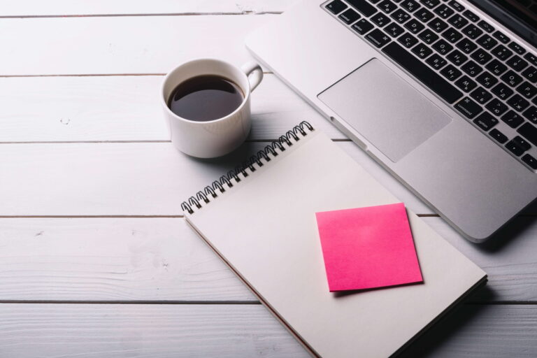 imagem de uma xícara branca com café preto, em cima de uma mesa e entre uma caderneta aberta com um post it rosa em cima e um notebook