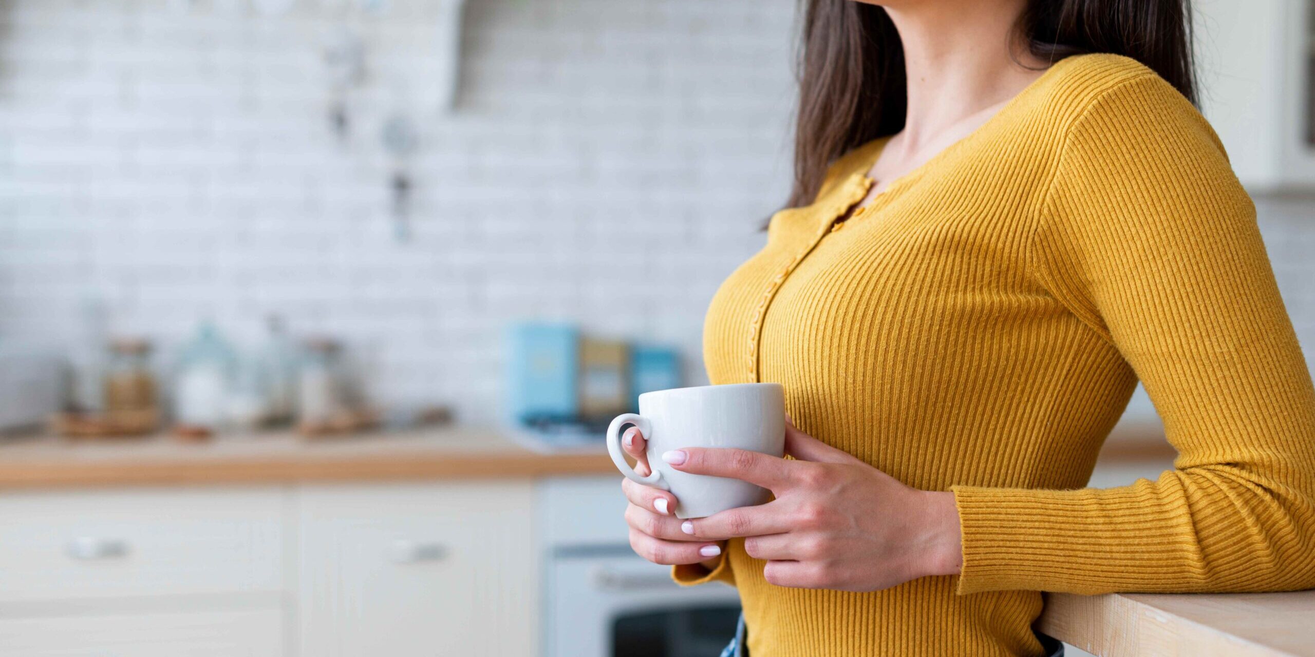 imagem de uma moça de cabelos longos castanhos, vestindo uma blusa de manga longa amarela e jeans, segurando uma xícara branca em um ambiente que parece ser uma cozinha em alusão a café ajuda a emagrecer