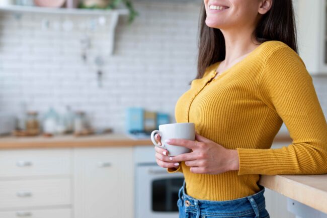 imagem de uma moça de cabelos longos castanhos, vestindo uma blusa de manga longa amarela e jeans, segurando uma xícara branca em um ambiente que parece ser uma cozinha em alusão a café ajuda a emagrecer