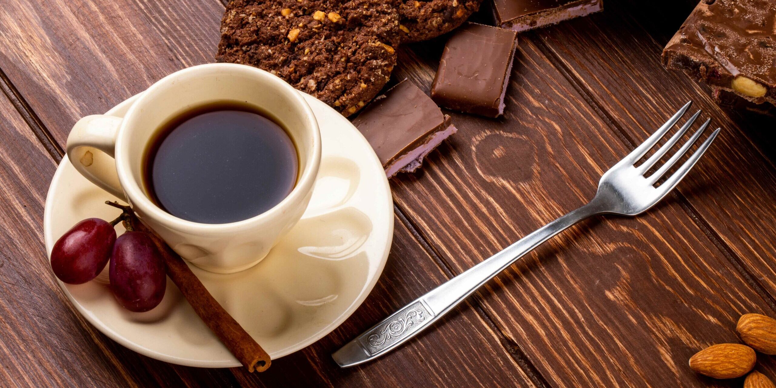 imagem de uma xícara com café e canela em pau ao lado e em volta um garfo, cookies e pedaços de chocolate em alusão a café doce ou café amargo