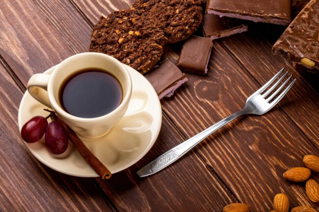 imagem de uma xícara com café e canela em pau ao lado e em volta um garfo, cookies e pedaços de chocolate em alusão a café doce ou café amargo